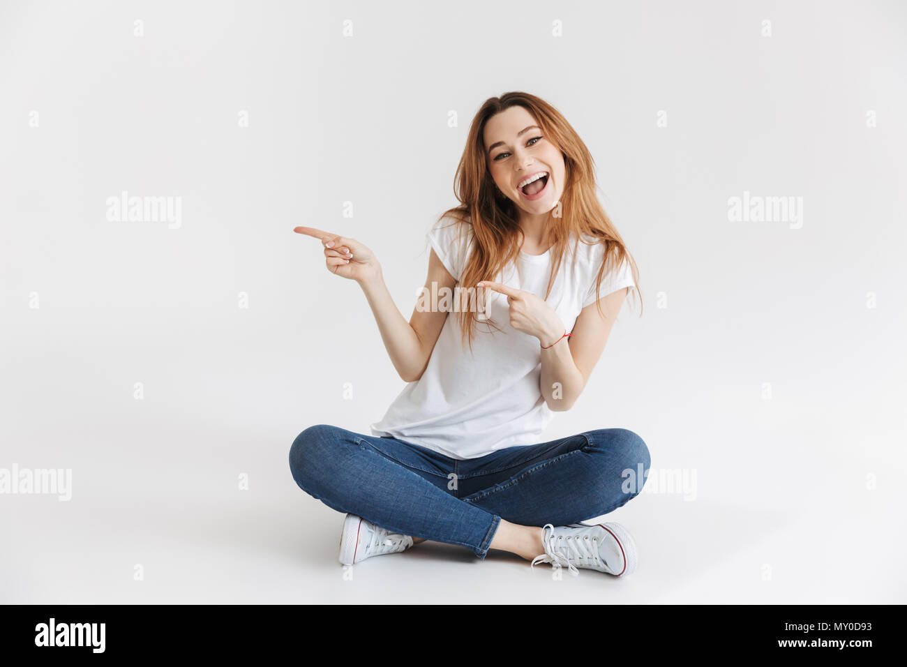 Happy woman in t-shirt sitting on the floor pointing away while looking ...