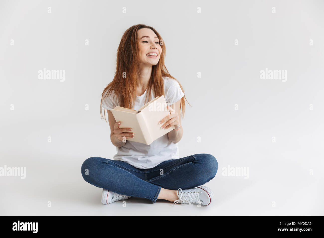 Portrait of a smiling young girl holding book while sitting with legs ...