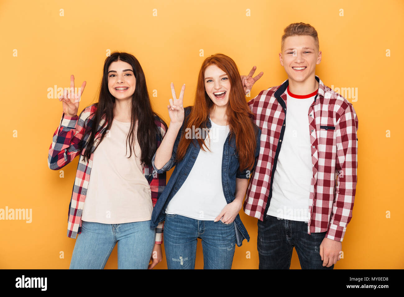 Group of happy school friends showing peace gesture while standing together over yellow background Stock Photo