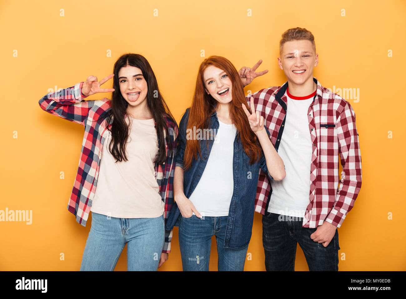 Group of happy school friends showing peace gesture while standing together and having fun over yellow background Stock Photo