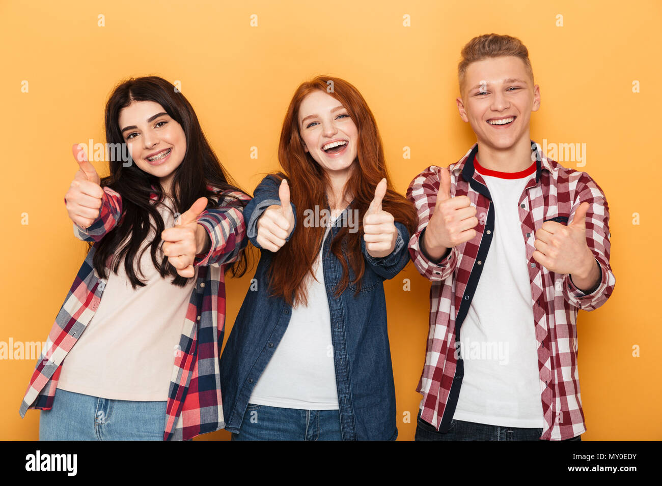 Group of cheerful school friends showing thumbs up while standing together over yellow background Stock Photo