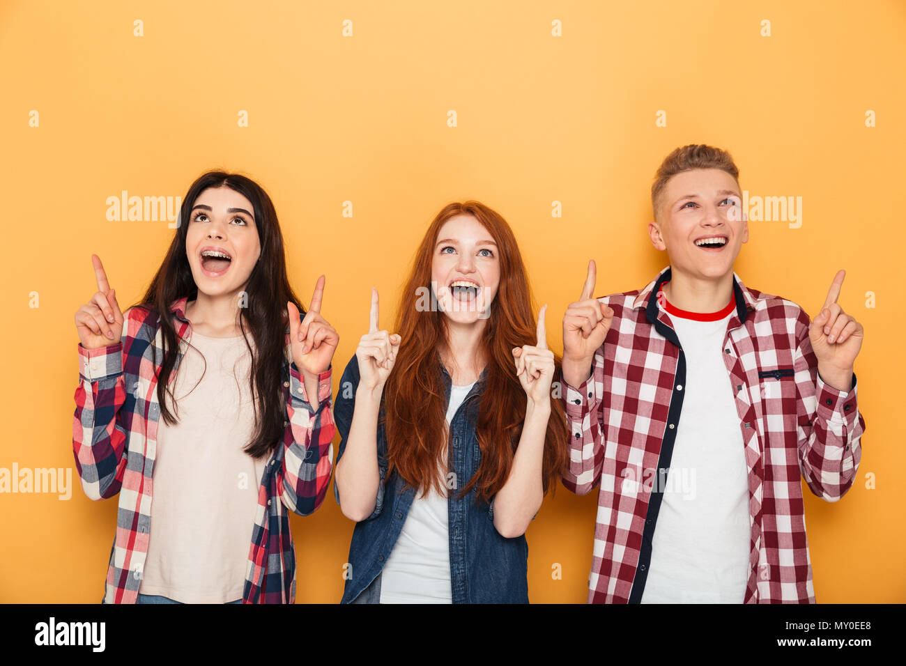 Group of happy school friends pointing fingers up at copy space while standing together over yellow background Stock Photo