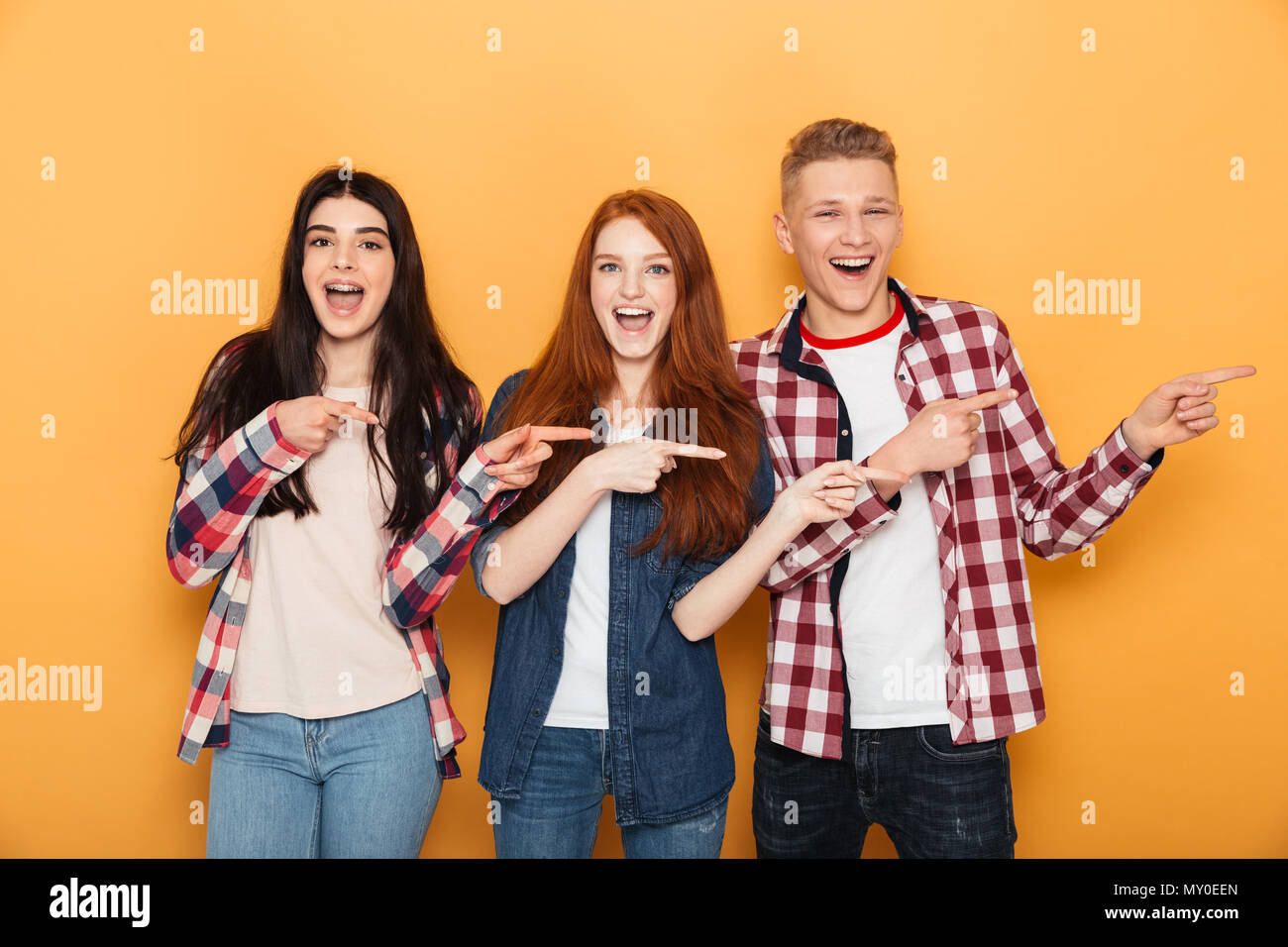 Group of cheerful school friends pointing fingers away at copy space while standing together over yellow background Stock Photo