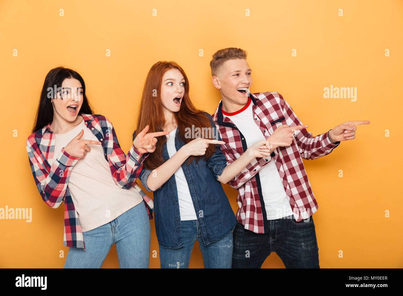 Group of an excited school friends pointing fingers away at copy space while standing together over yellow background Stock Photo
