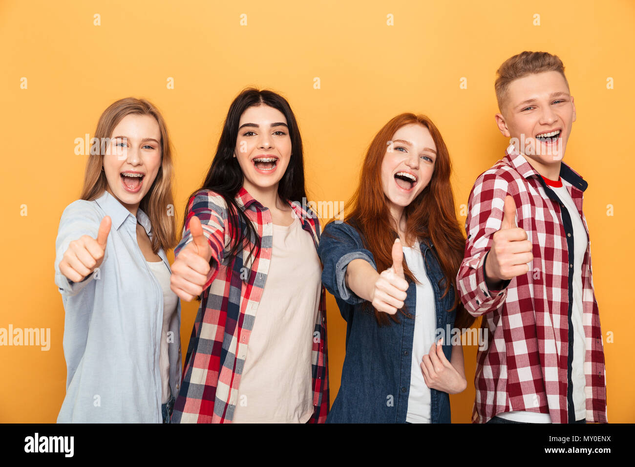 Group of smiling school friends showing thumbs up while standing together over yellow background Stock Photo