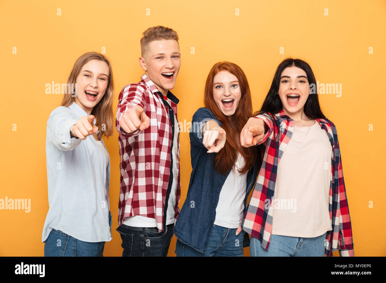 Group of happy school friends pointing fingers at camera while standing together over yellow background Stock Photo