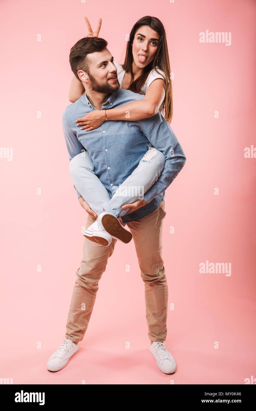 Full length portrait of a happy young man giving his girlfriend piggyback ride and having fun isolated over pink background Stock Photo image