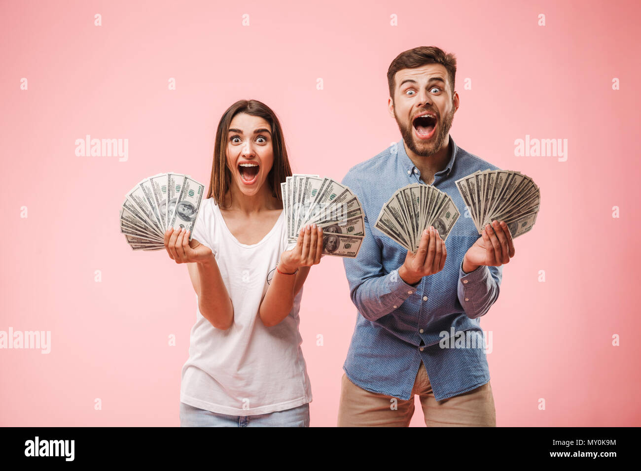 Portrait of a joyful young couple holding bunch of money banknotes and celebrating success isolated over pink background Stock Photo