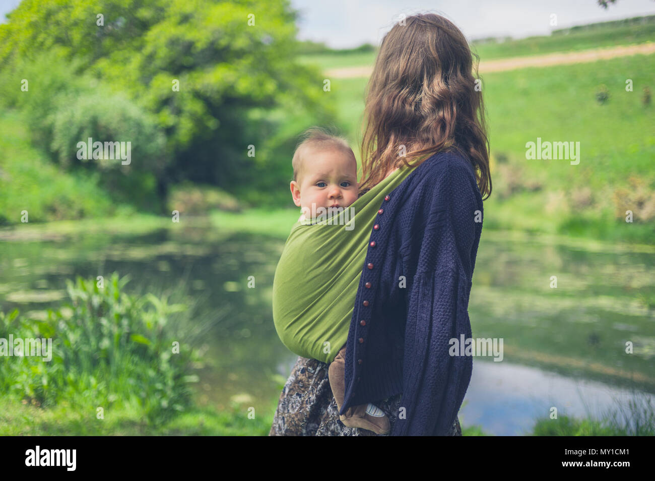 A young woman with a baby in a sling is standing by a pond in the countryside Stock Photo