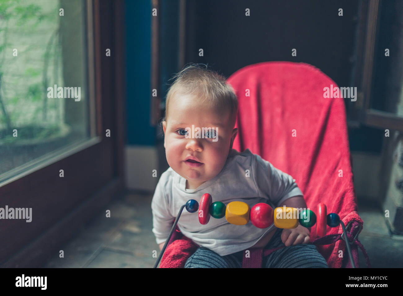 A little baby is sitting in a bouncy chair by the window Stock Photo