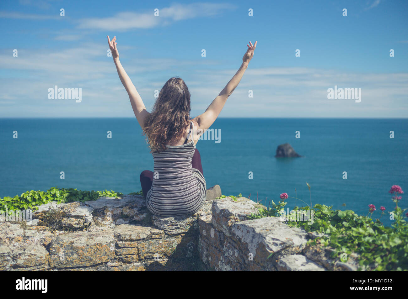 A young woman is sitting on a wall by the sea and is expressing freedom ...