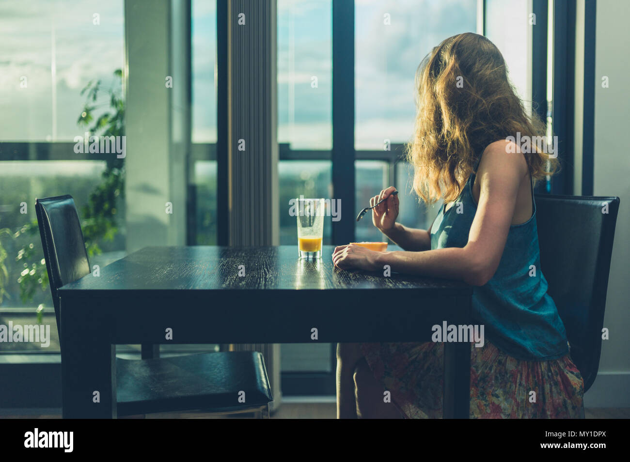 A young woman is sitting at a table by the window in a modern city apartment and is having breakfast Stock Photo