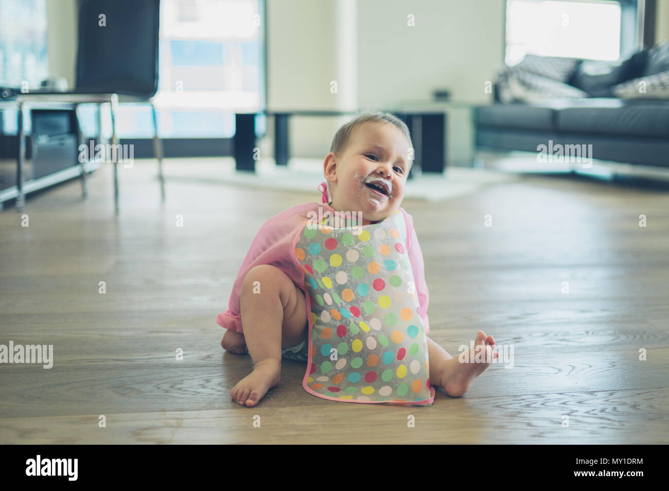 A cute little baby wearing a bib is sitting on the kitchen floor with yogurt on his face Stock Photo