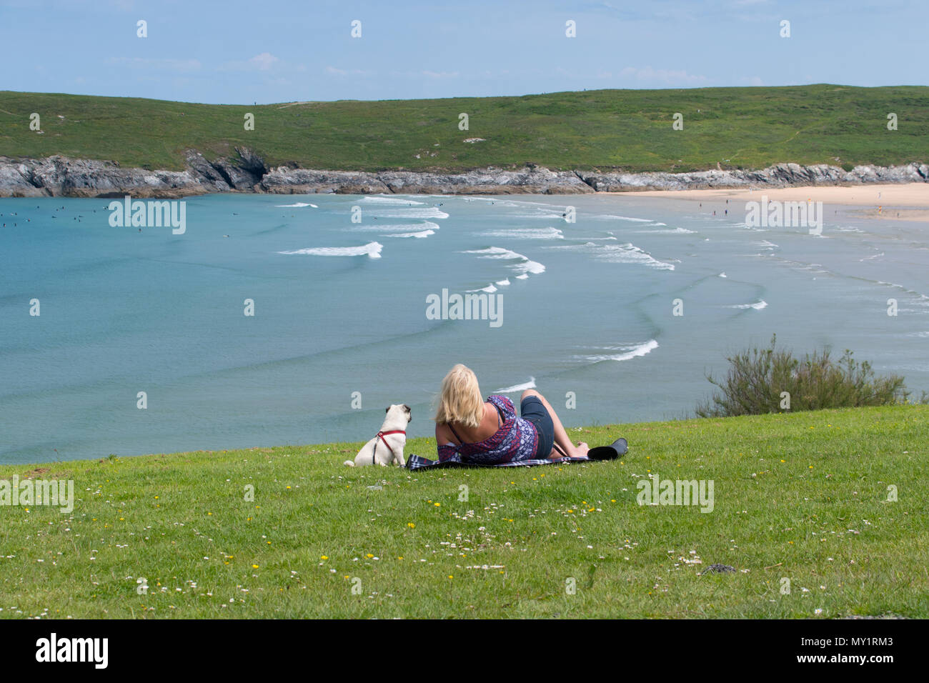 Woman and her dog sitting / lying on the grass looking towards the sea and Crantock beach in Cornwall Stock Photo