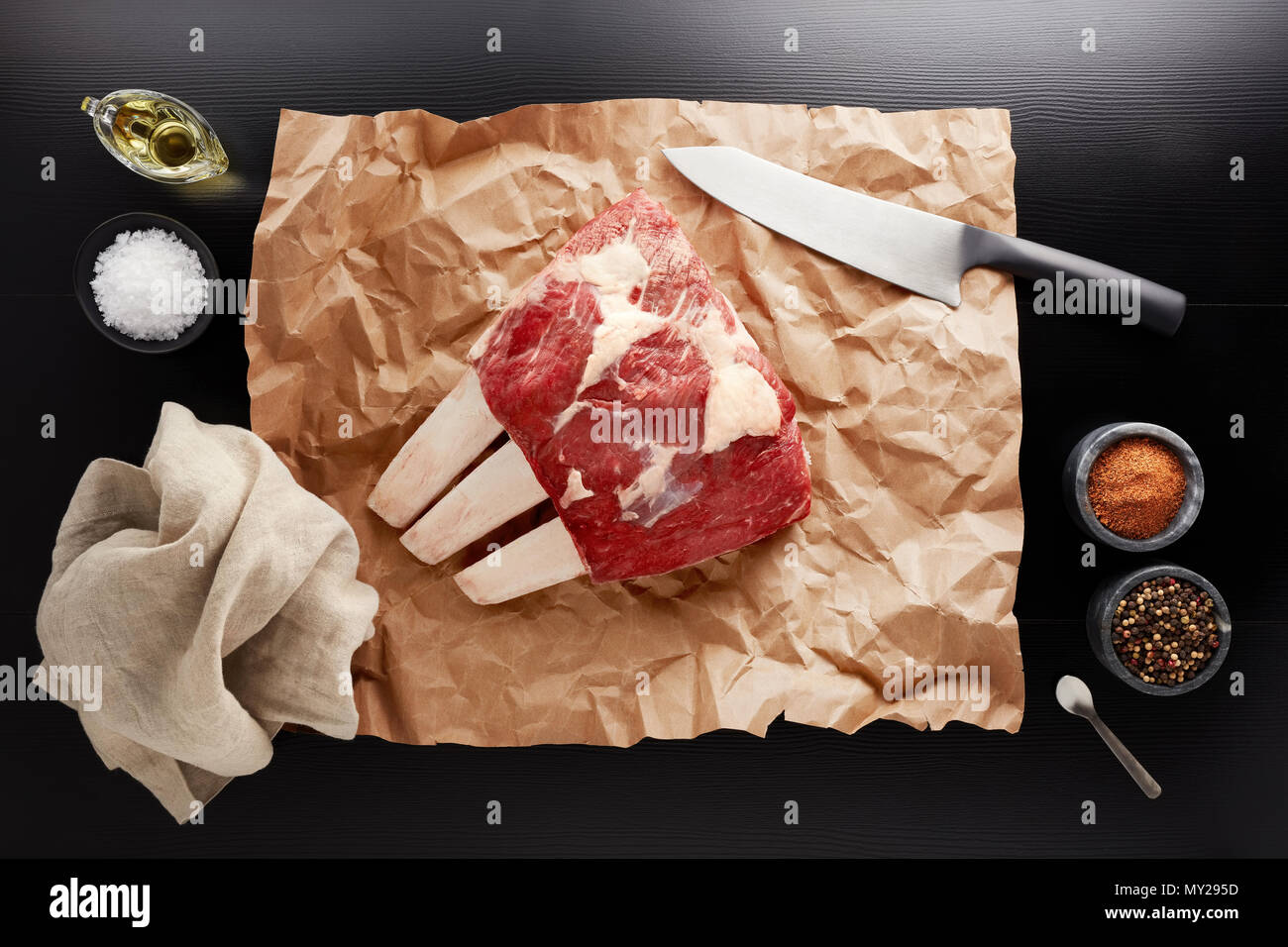 Raw uncooked veal rib. Top down view still life image with knife, paper bag, spices, napkin, salt and oil on a black table. Stock Photo