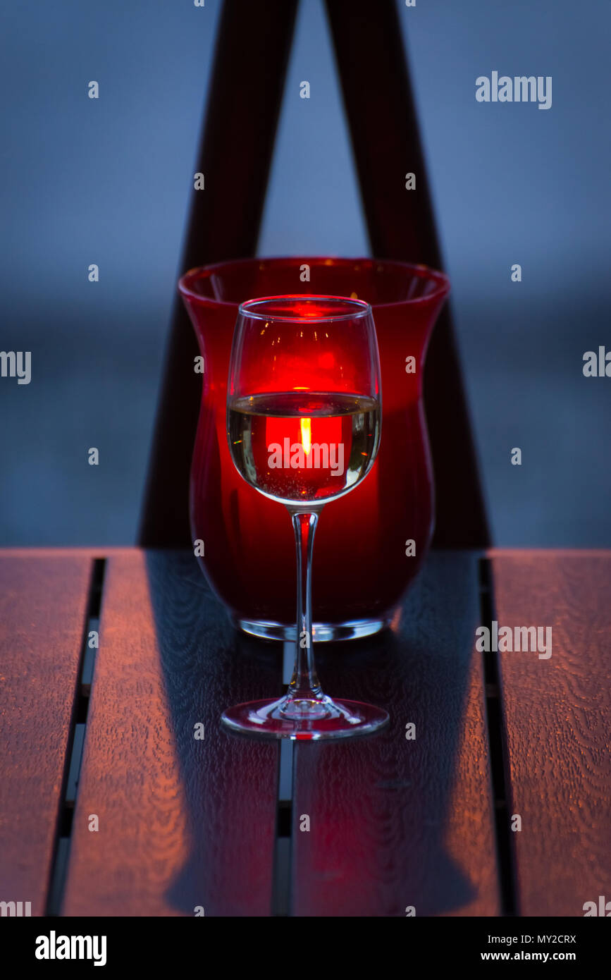 A glass of wine is on the table, and behind it a candle is burning in a red vase Stock Photo