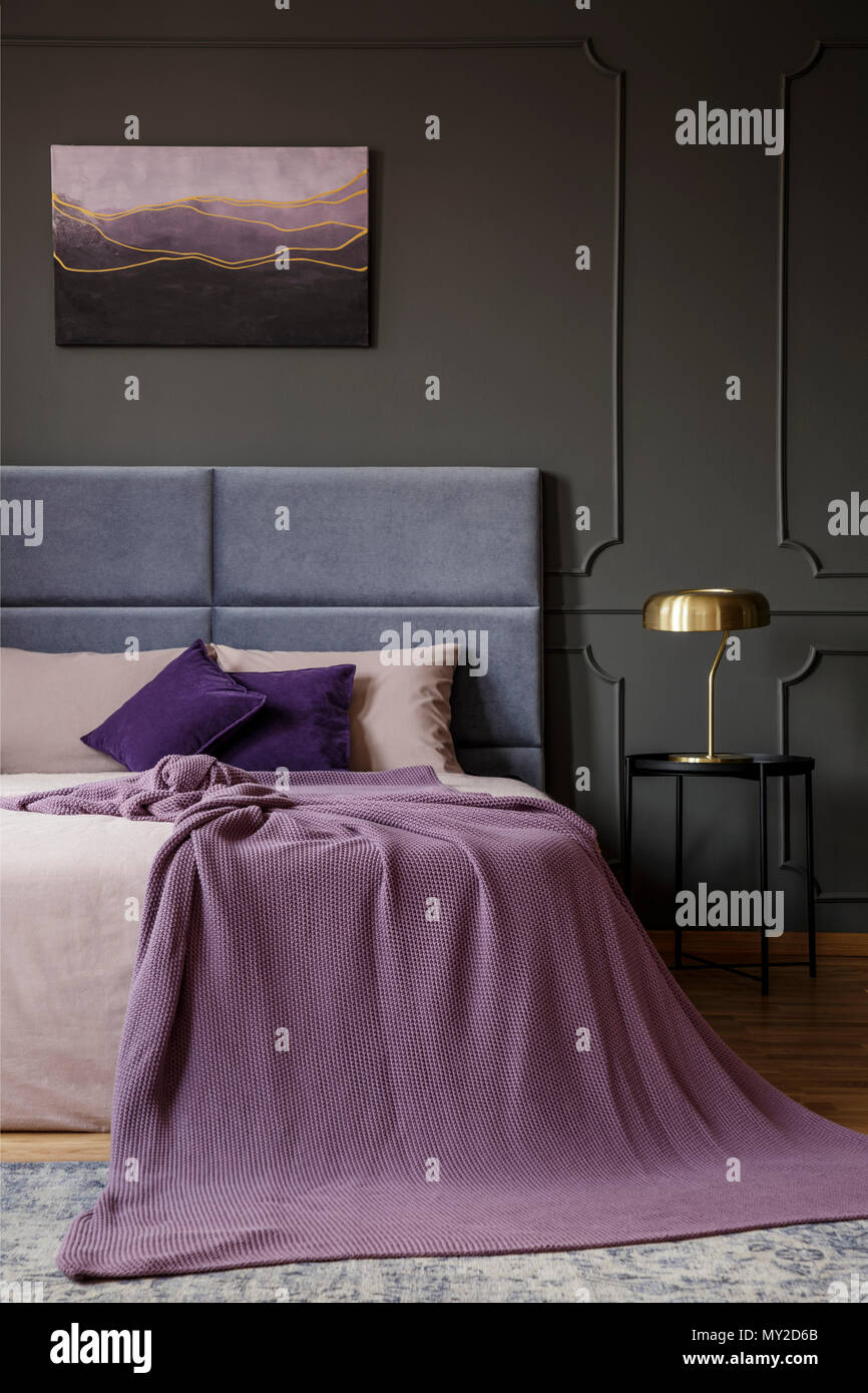 Gold lamp on black table next to bed with pink blanket in pastel bedroom interior with poster Stock Photo