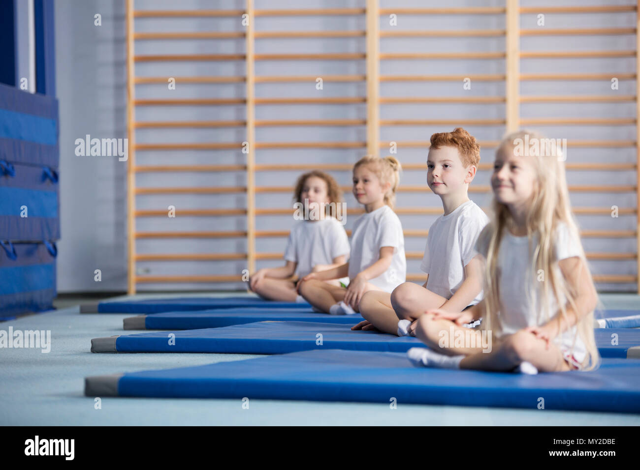 Smiling pupils sitting on blue mats during corrective gymnastics classes Stock Photo