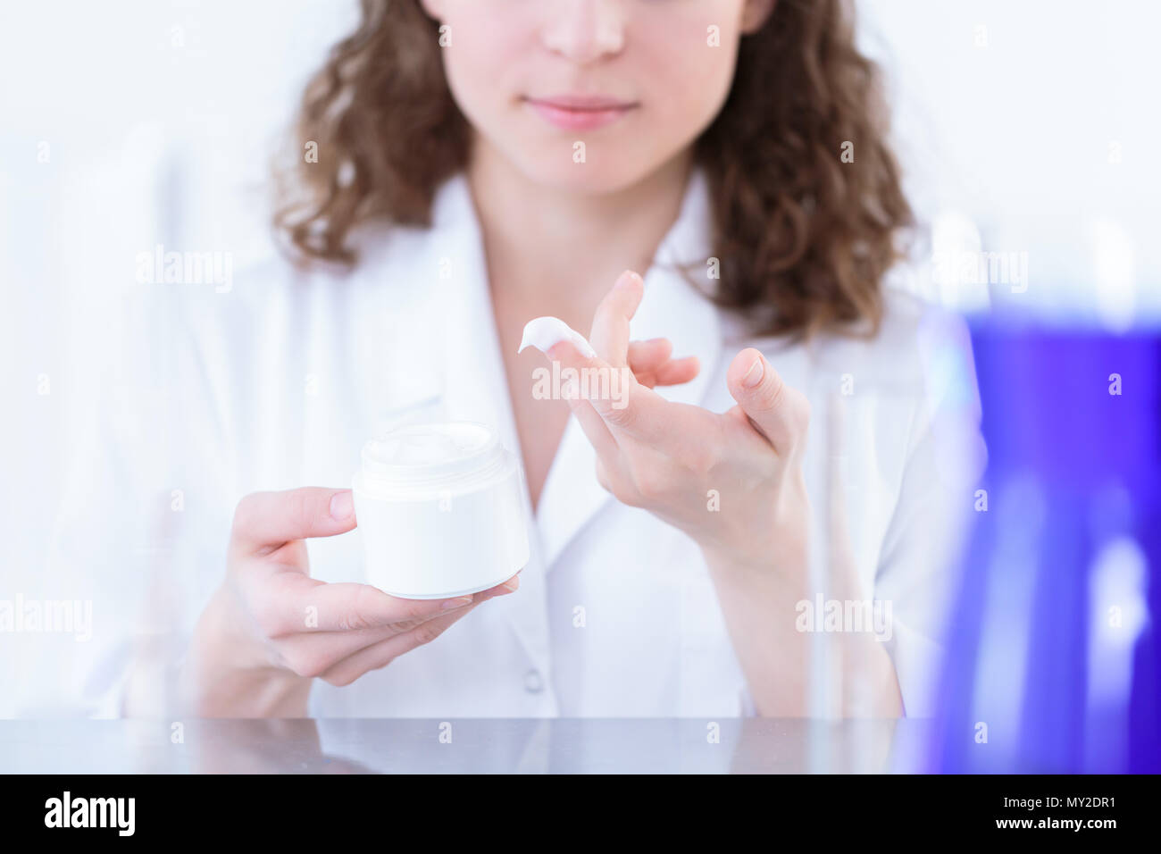 Female chemist in laboratory coat holding a white container and presenting lotion on her finger Stock Photo