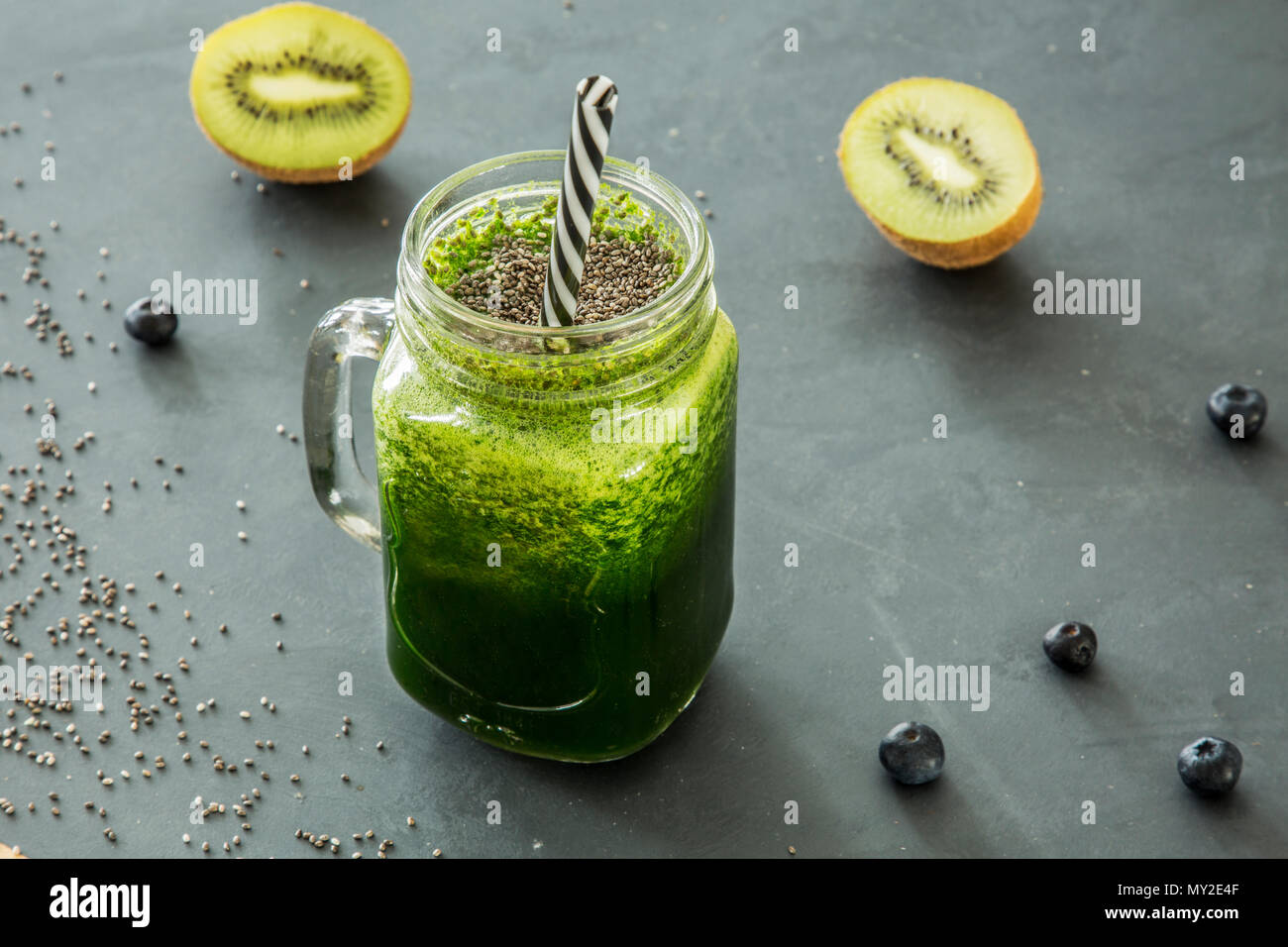 Green, healthy smoothie in a glass jar with a straw and kiwi, blueberries and chia seeds scattered around Stock Photo