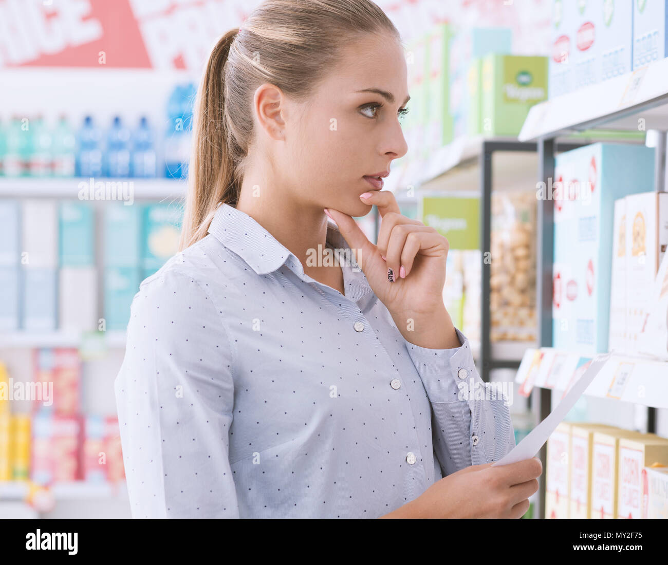 Young woman at the supermarket, she is holding a shopping list and searching products on a shelf Stock Photo