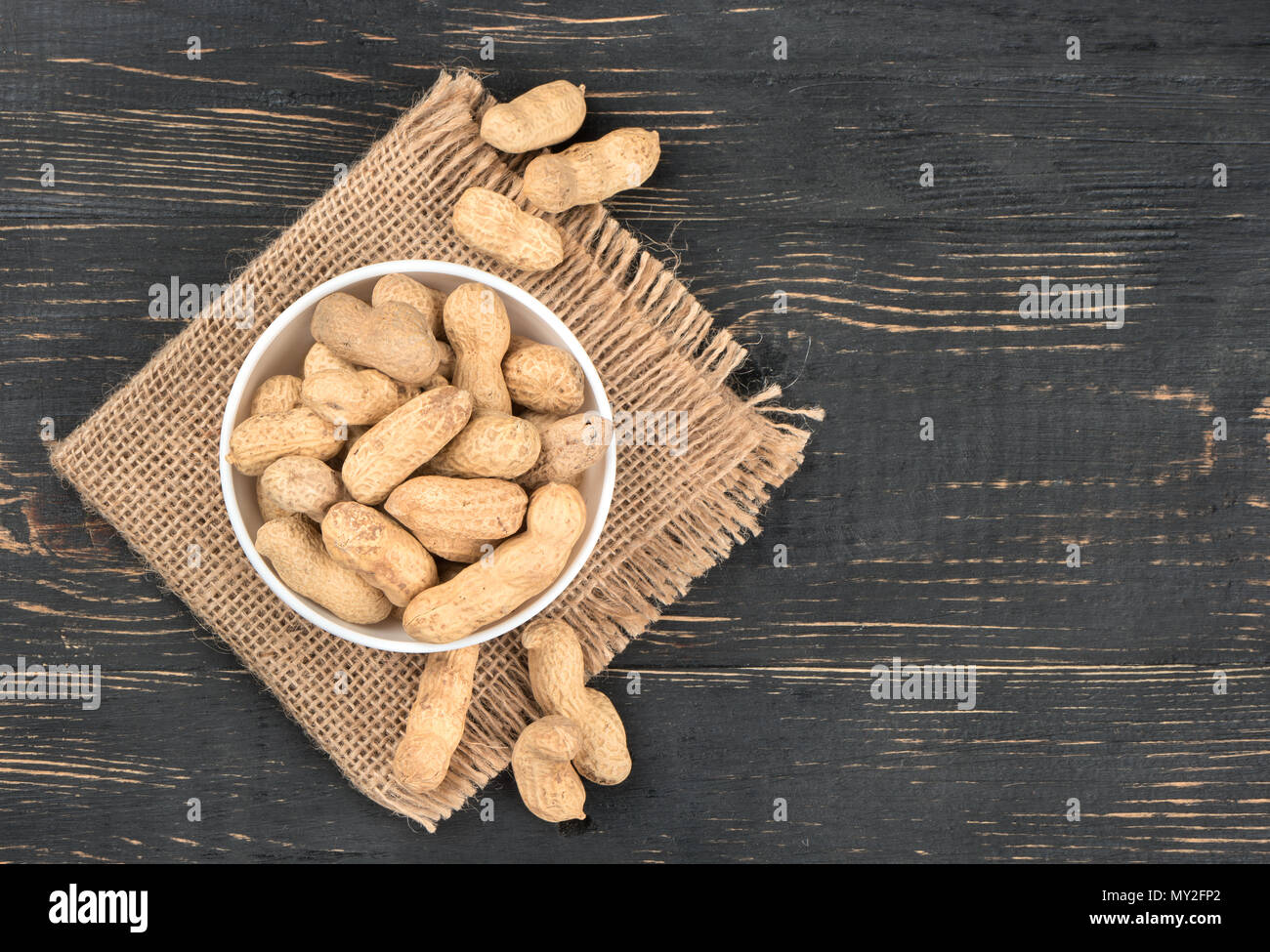 Full ceramic bowl of peanuts in the shell on sackcloth and empty wooden background, top view Stock Photo