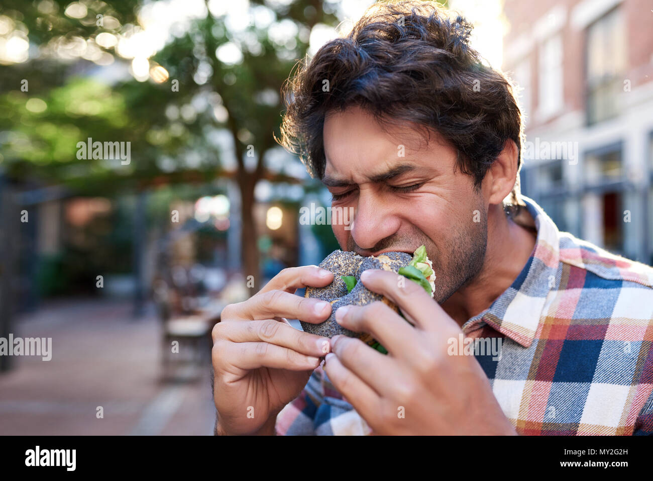 Hungry young man standing outside with his eyes closed and taking a bite out of his delicious poppy seed bagel Stock Photo