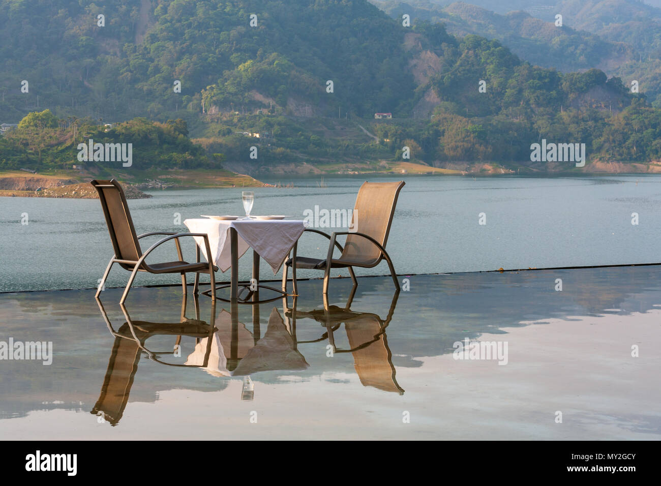 Table and chairs in water reflected in infinity pool overlooking lake and mountain landscape Stock Photo