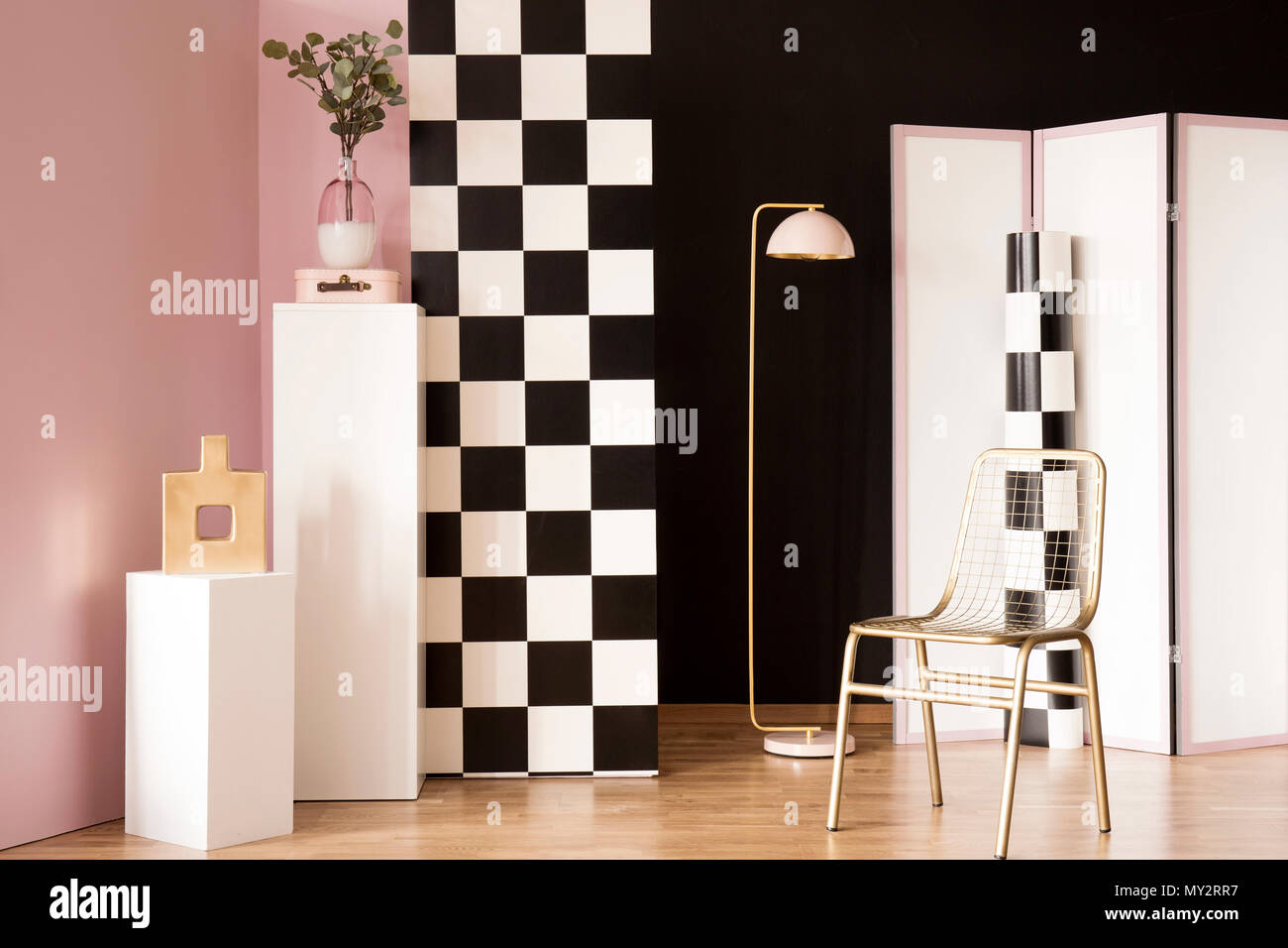 Gold chair next to pink lamp in pastel interior with checkerboard wall and plant on white pedestal Stock Photo