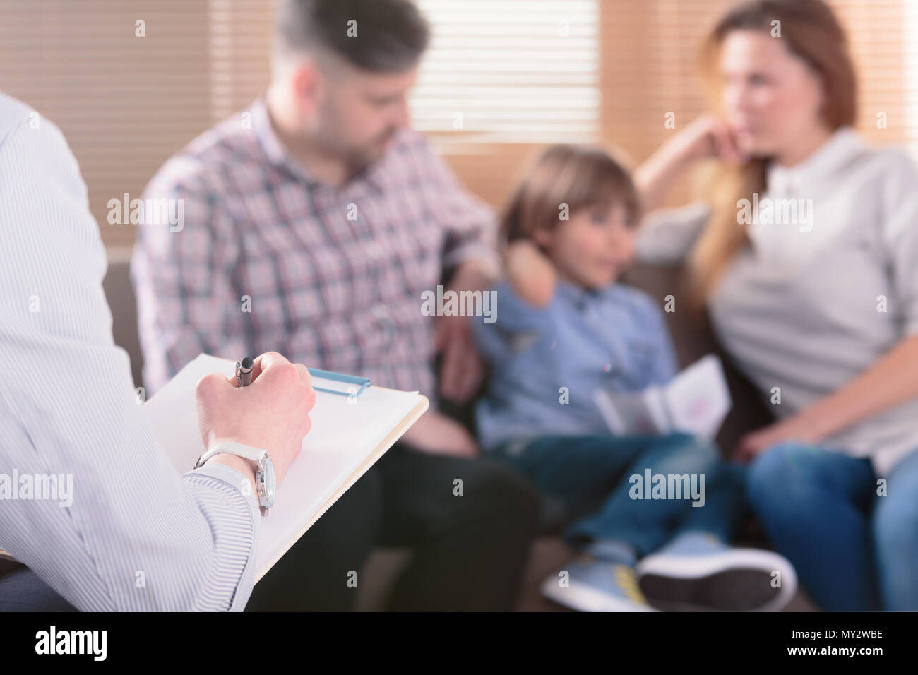 Hand of a professional family psychotherapist writing notes in front of a couple with a child in a blurred background during a consultation Stock Photo