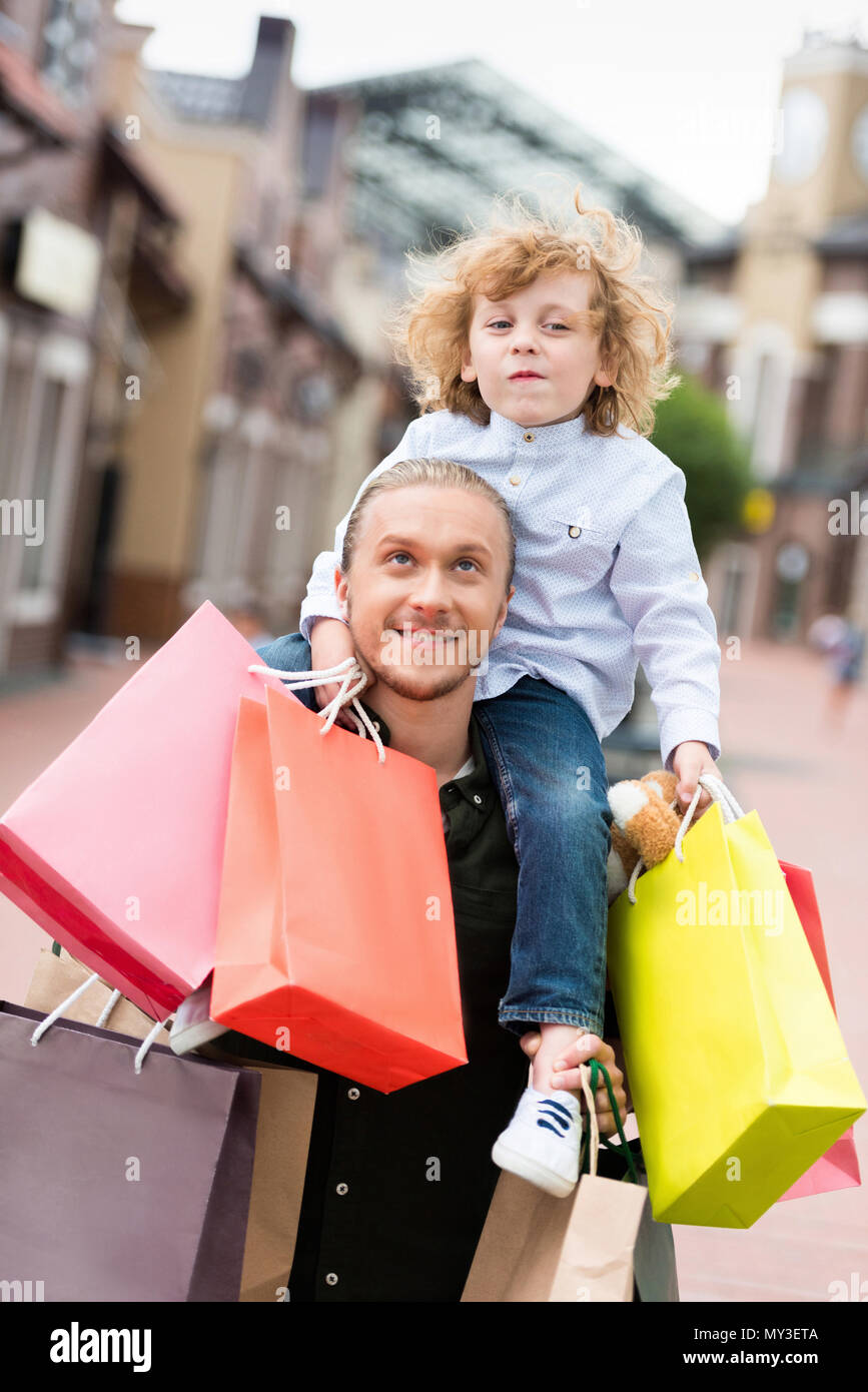 portrait of smiling father piggybacking his little son with shopping bags at street Stock Photo