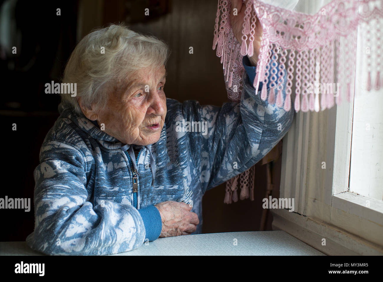 An elderly woman in the house sitting at the table looking out the window. Age 90 years old. Stock Photo