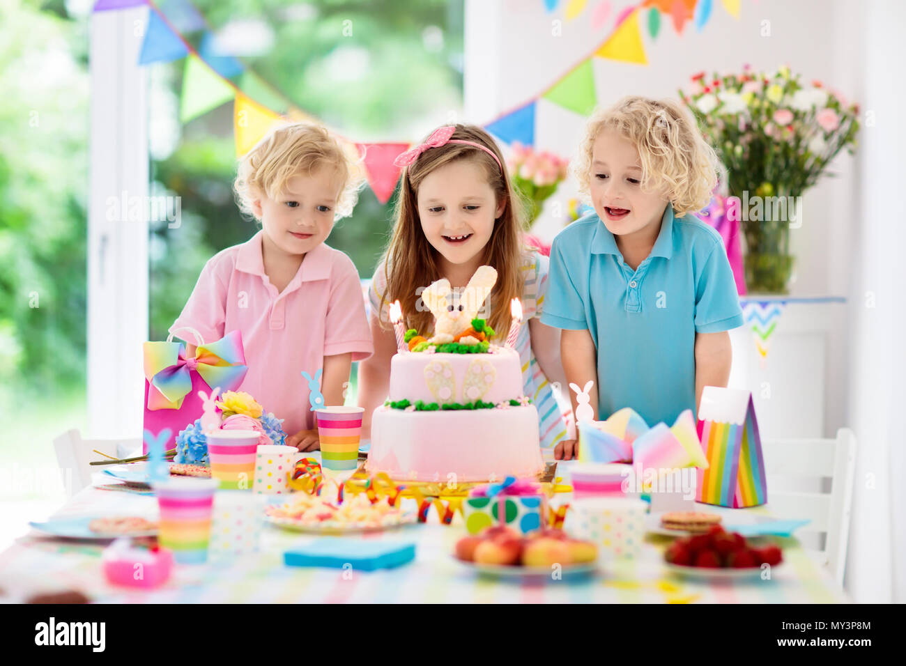 Kids birthday party. Children blow out candles on pink bunny cake. Pastel rainbow decoration and table setting for kids event, banner and flag. Girl a Stock Photo