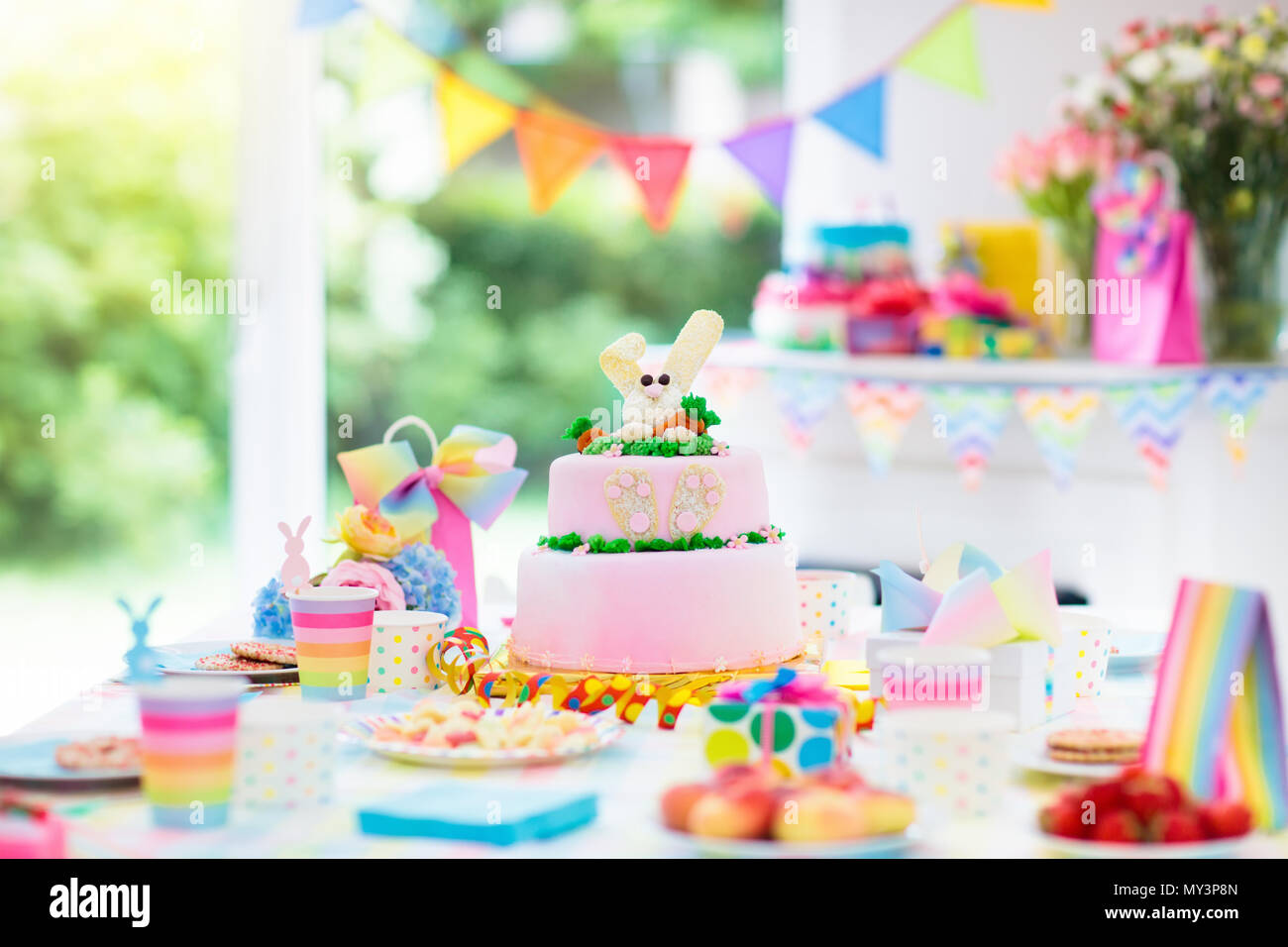 Kids birthday party decoration and cake. Decorated table for child birthday celebration. Rainbow bunny cake for little girl. Room with festive balloon Stock Photo