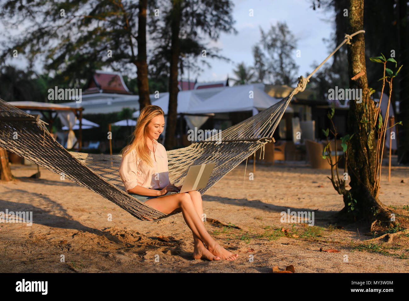 Young woman using laptop and sitting on sand in wicker hammock. Stock Photo