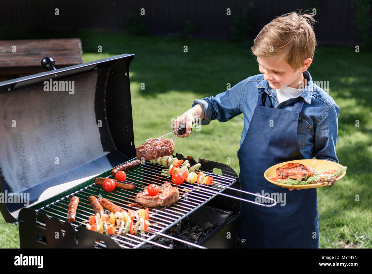 smiling kid boy in apron preparing tasty stakes on barbecue grill outdoors  Stock Photo - Alamy