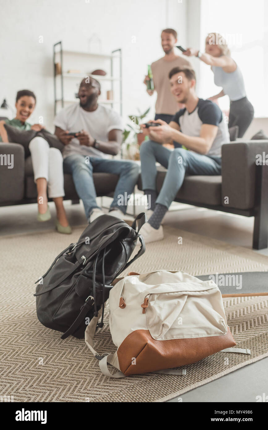 two backpacks and longboard on foreground and group of multiethnic friends with joysticks playing video game Stock Photo