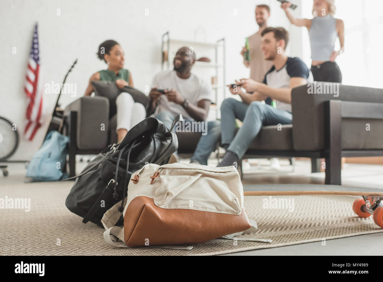 closeup shot of two backpacks and longboard on foreground and group of multiethnic friends with joysticks playing video game Stock Photo