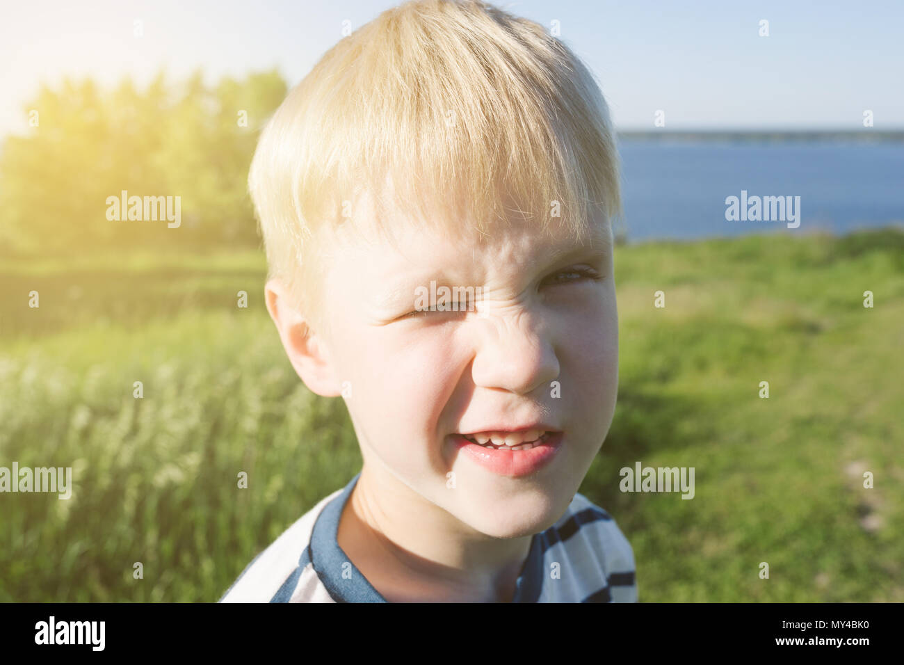 Portrait of blond boy joyful and closing his eye from the sun on nature. Summer activity. Stock Photo