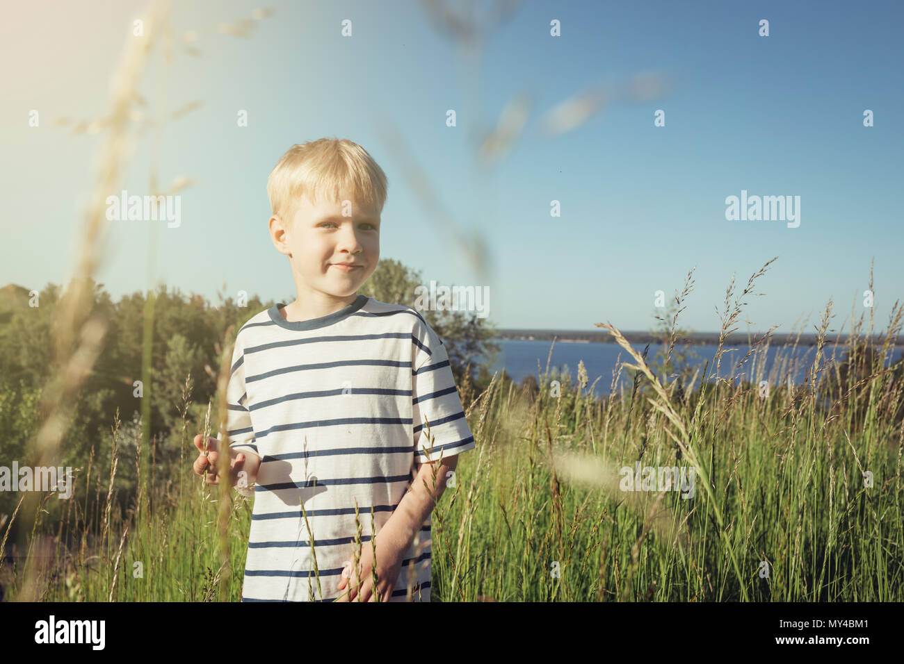 Little blond boy smiling and looking at camera on nature. Summer activity. Field with high grass. Stock Photo