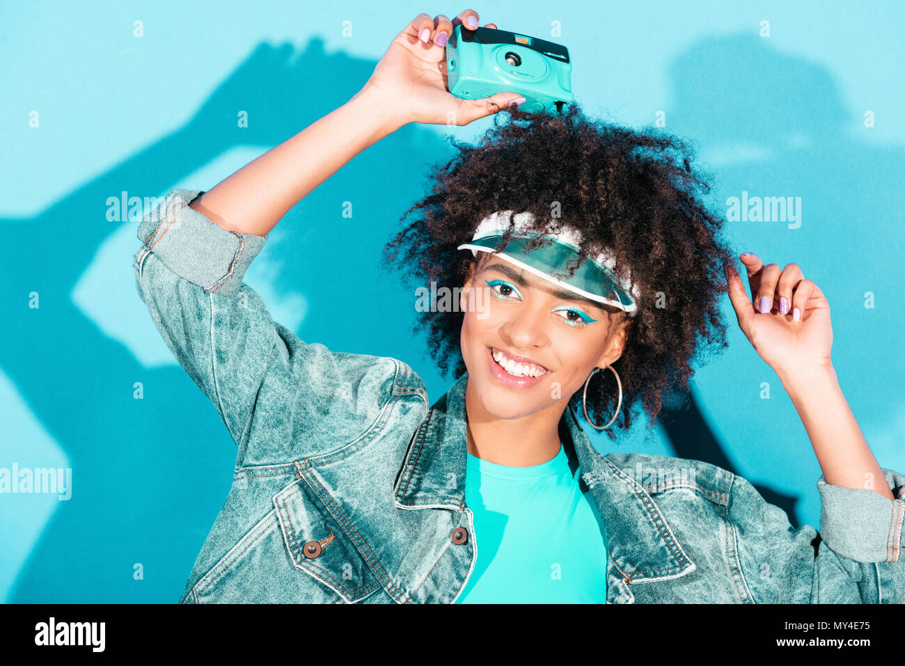 beautiful afro model posing in jeans jacket with vintage photo camera, on turquoise Stock Photo