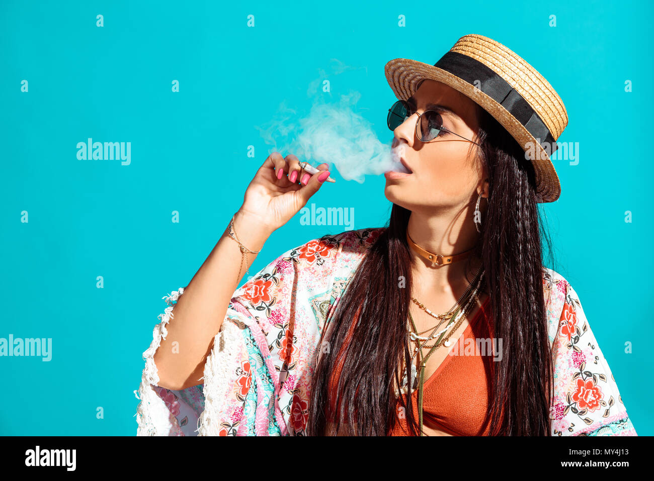 Attractive bohemian girl smoking cigarette and exhaling smoke isolated on turquoise Stock Photo