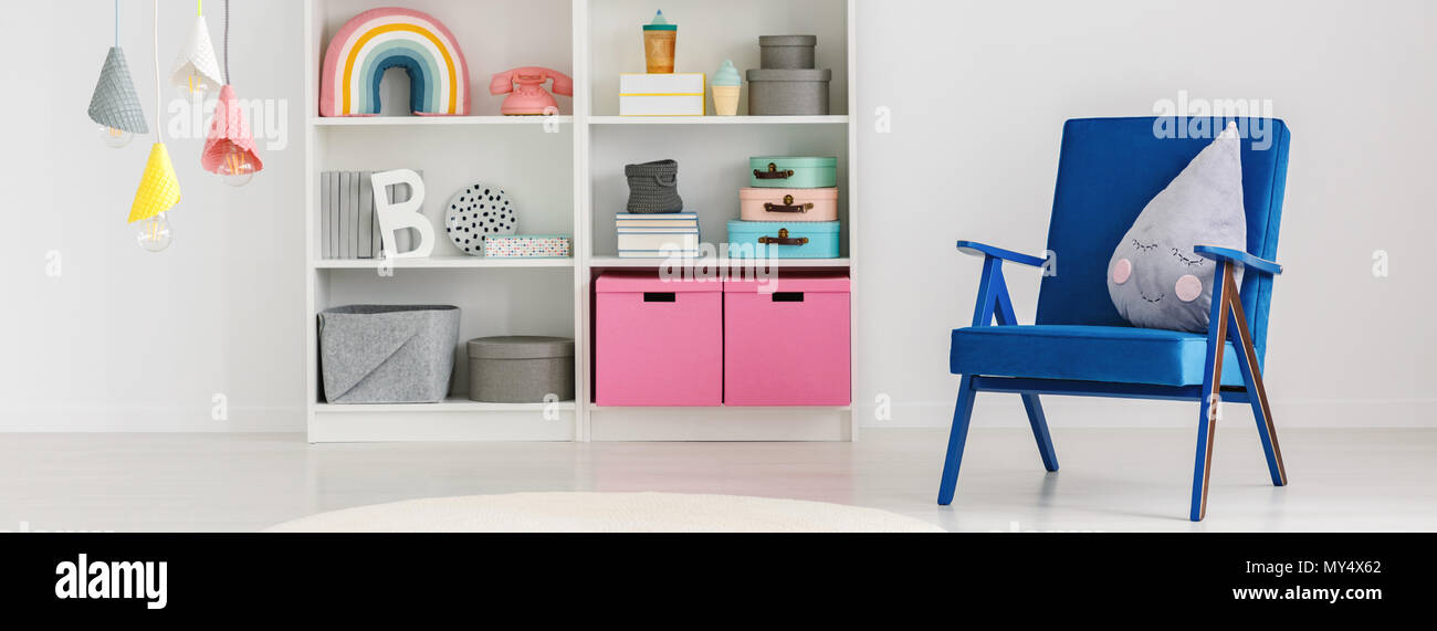 Panorama of a cute pillow on a blue armchair and a bookcase with boxes and decorations in a fun child bedroom interior with white walls Stock Photo