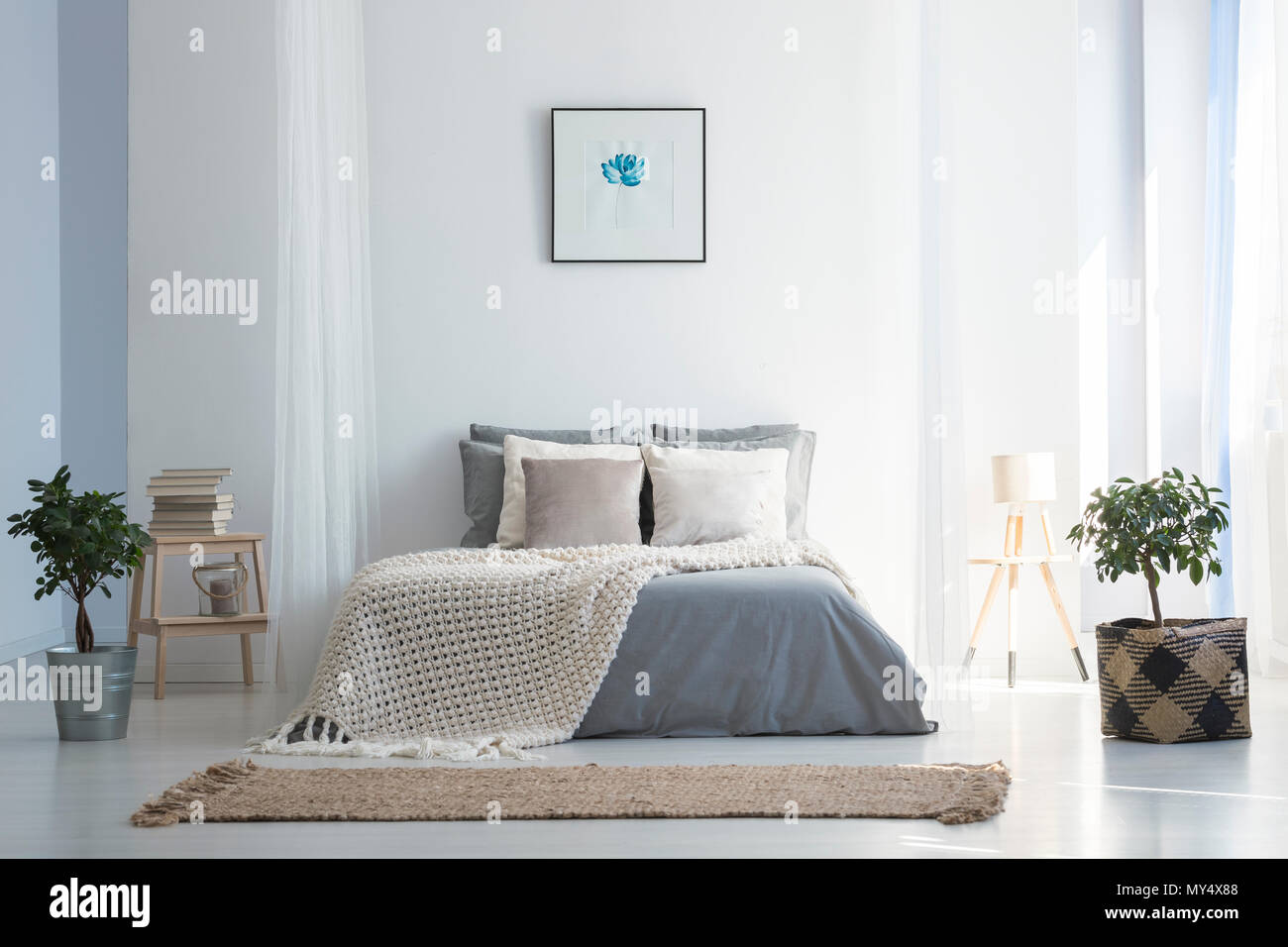 Knit blanket on grey bed in bright bedroom interior with poster and plants. Real photo Stock Photo