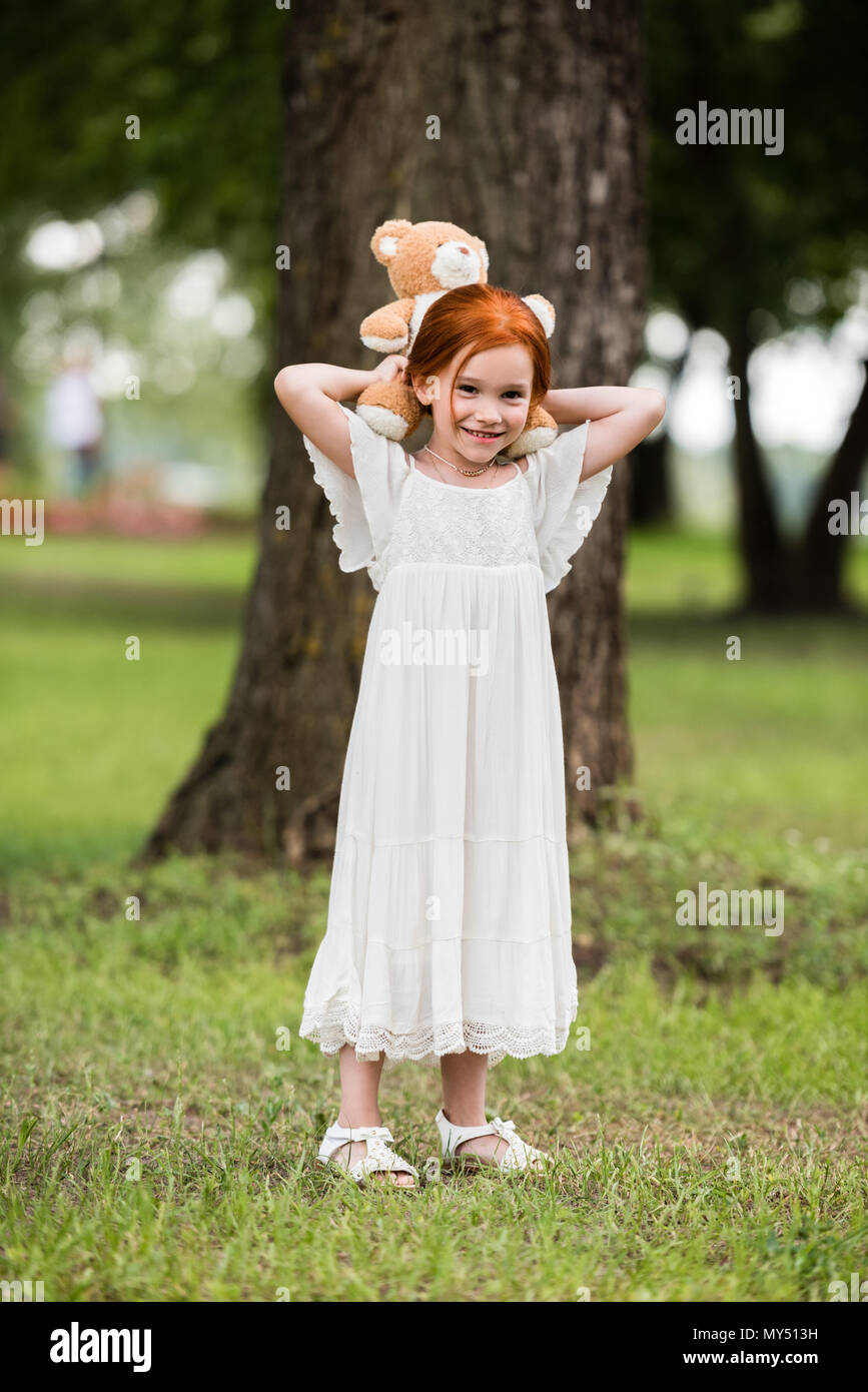 Cute Little Red Haired Girl In White Dress Holding Teddy Bear At
