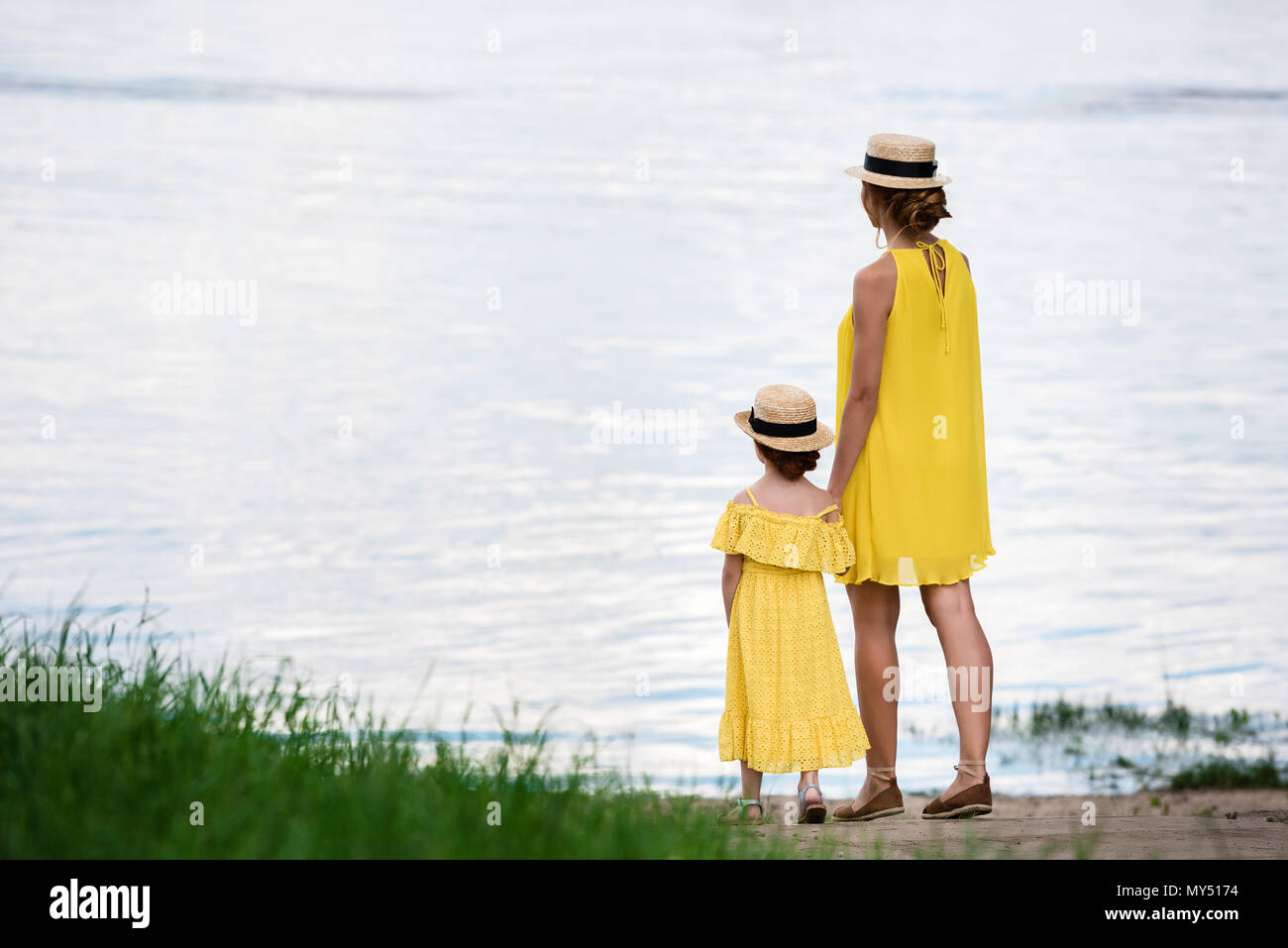 back view of pretty mother and daughter holding hands while standing at riverside Stock Photo