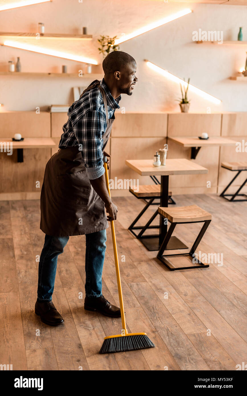handsome smiling african american worker sweeping with broom in coffee