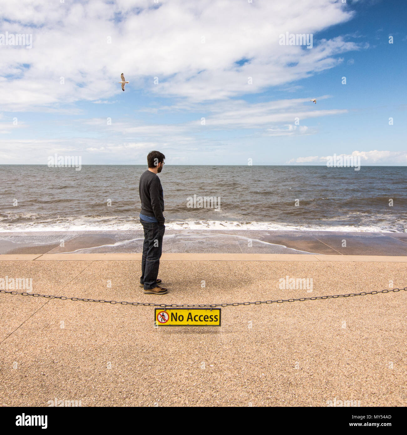Blackpool, England, UK - August 1, 2015: A man looks at the Blackpool Beach sea defences at high tide, behind a No Access sign, while gulls fly overhe Stock Photo