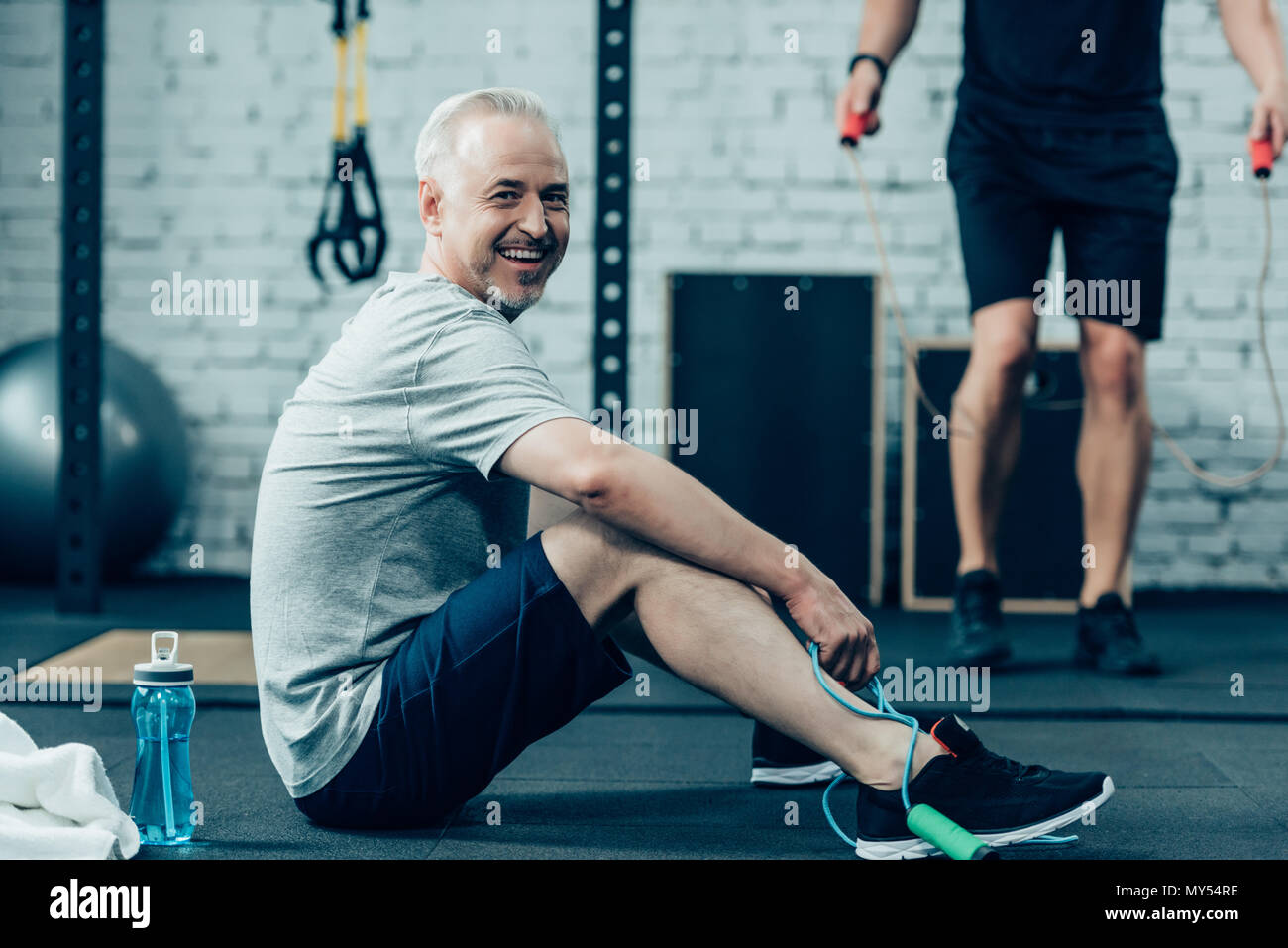 senior sportsman resting while young sportsman training with skipping rope in gym Stock Photo
