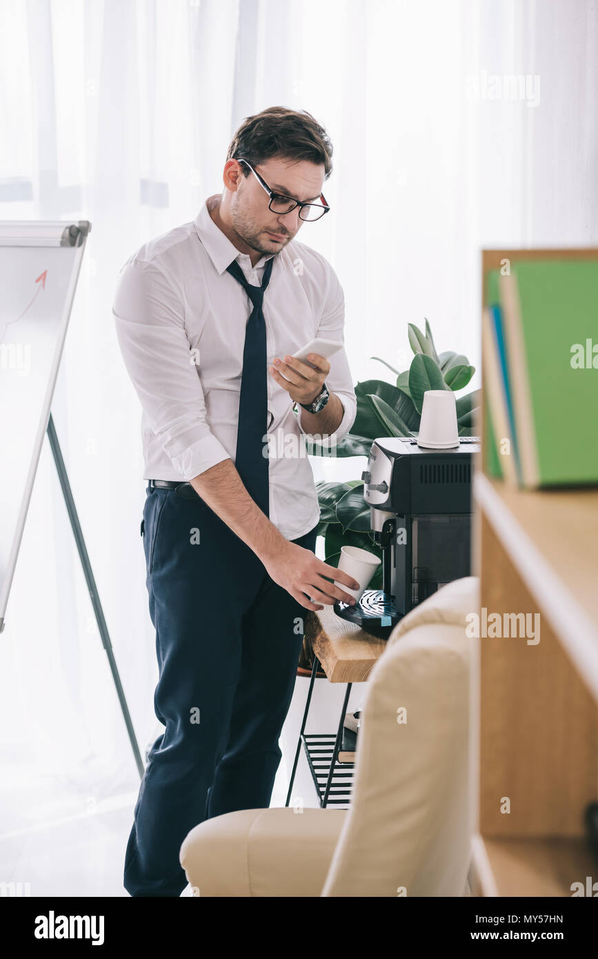 untidy businessman pouring coffee from office machine and using smartphone Stock Photo